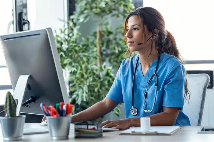 Healthcare professional in blue scrubs with stethoscope at a computer, wearing a headset for telemedicine or patient support.
